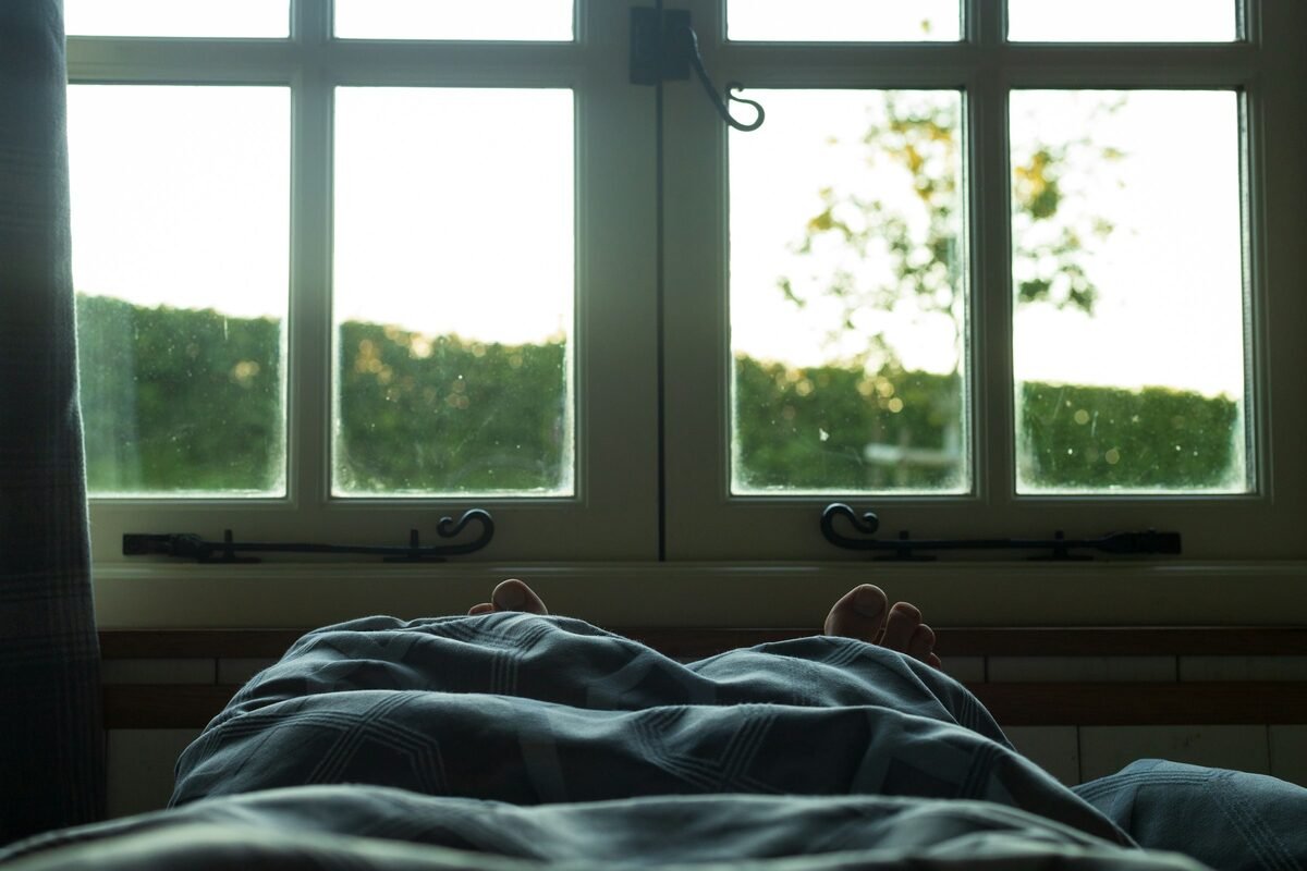 Deux pieds qui dépassent d'une couette bleu-gris, sur un lit placé devant une fenêtre aux petits carreaux