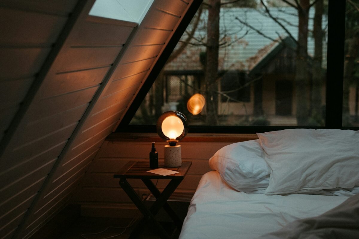 Chambre mansardée le soir, une lampe ronde allumée sur une petite table à côté d'un lit aux oreillers blancs