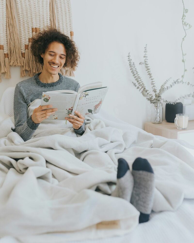 Homme souriant qui lit un livre dans son lit sous un plaid clair, les pieds en chaussettes grises et bleu marine au premier plan